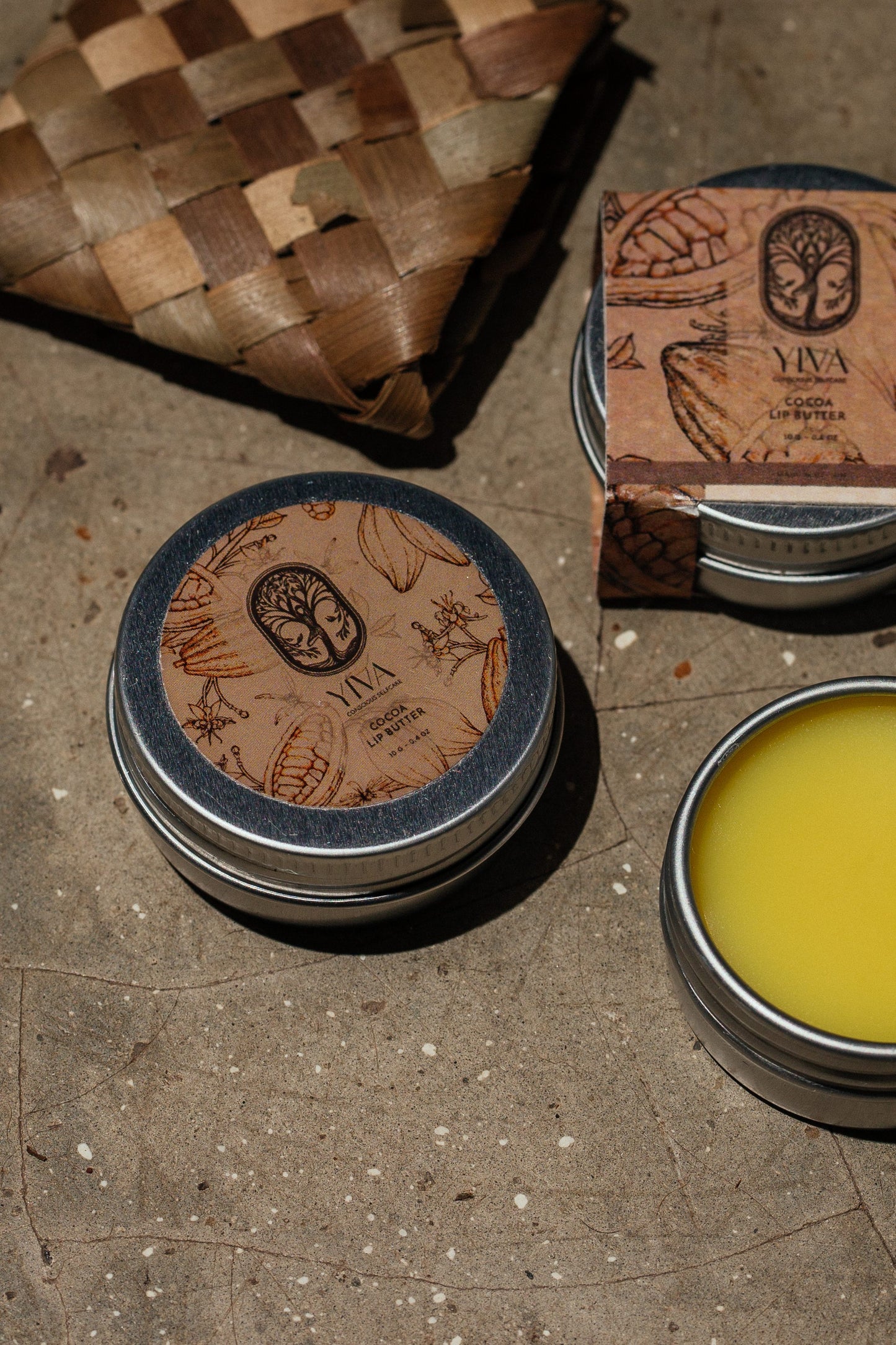Two tins of Yiva Ceylon lipbalm on a textured surface with a woven basket in the background.