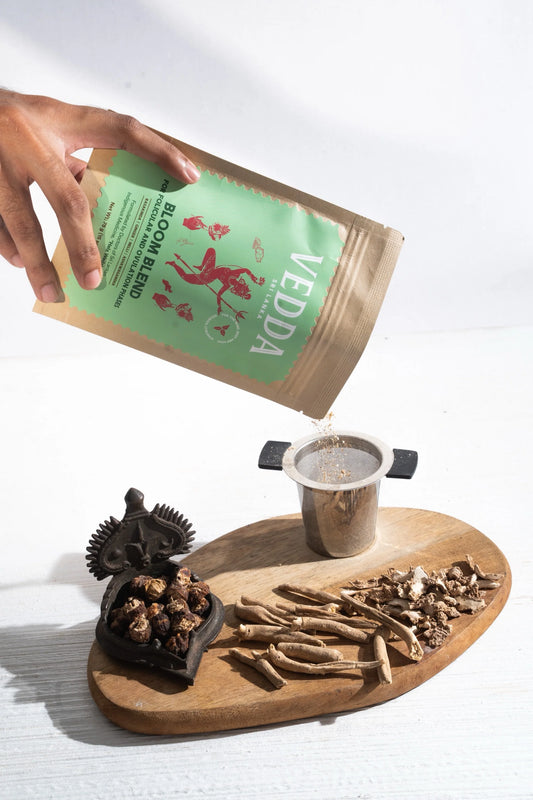 Hand pouring Vedda tea into a cup on a wooden tray with tea leaves and a tea bag.