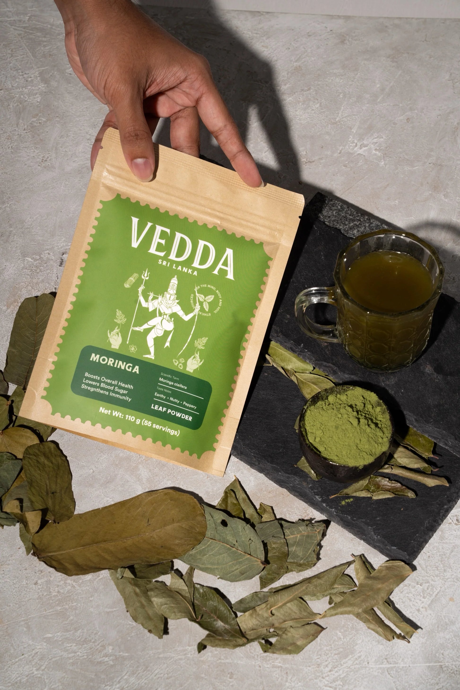 Vedda Moringa leaf powder package with moringa leaves and a cup of tea on a stone surface
