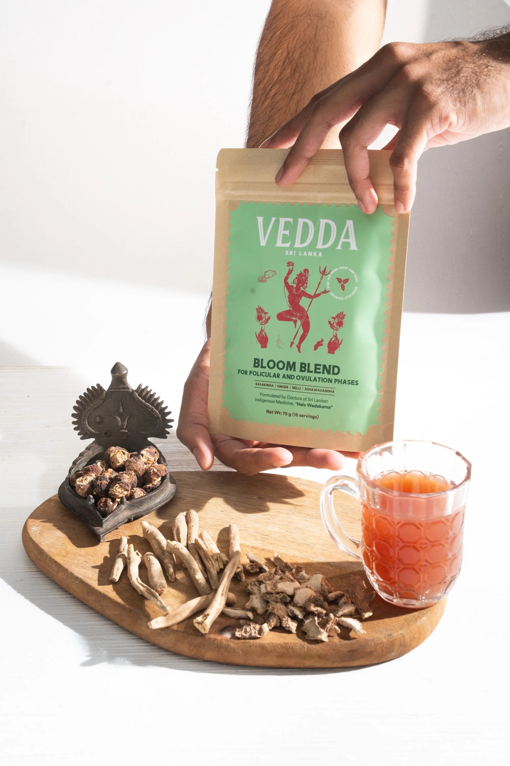 Vedda Bloom Blend package held by a hand with a wooden board featuring dried herbs and a glass of tea.