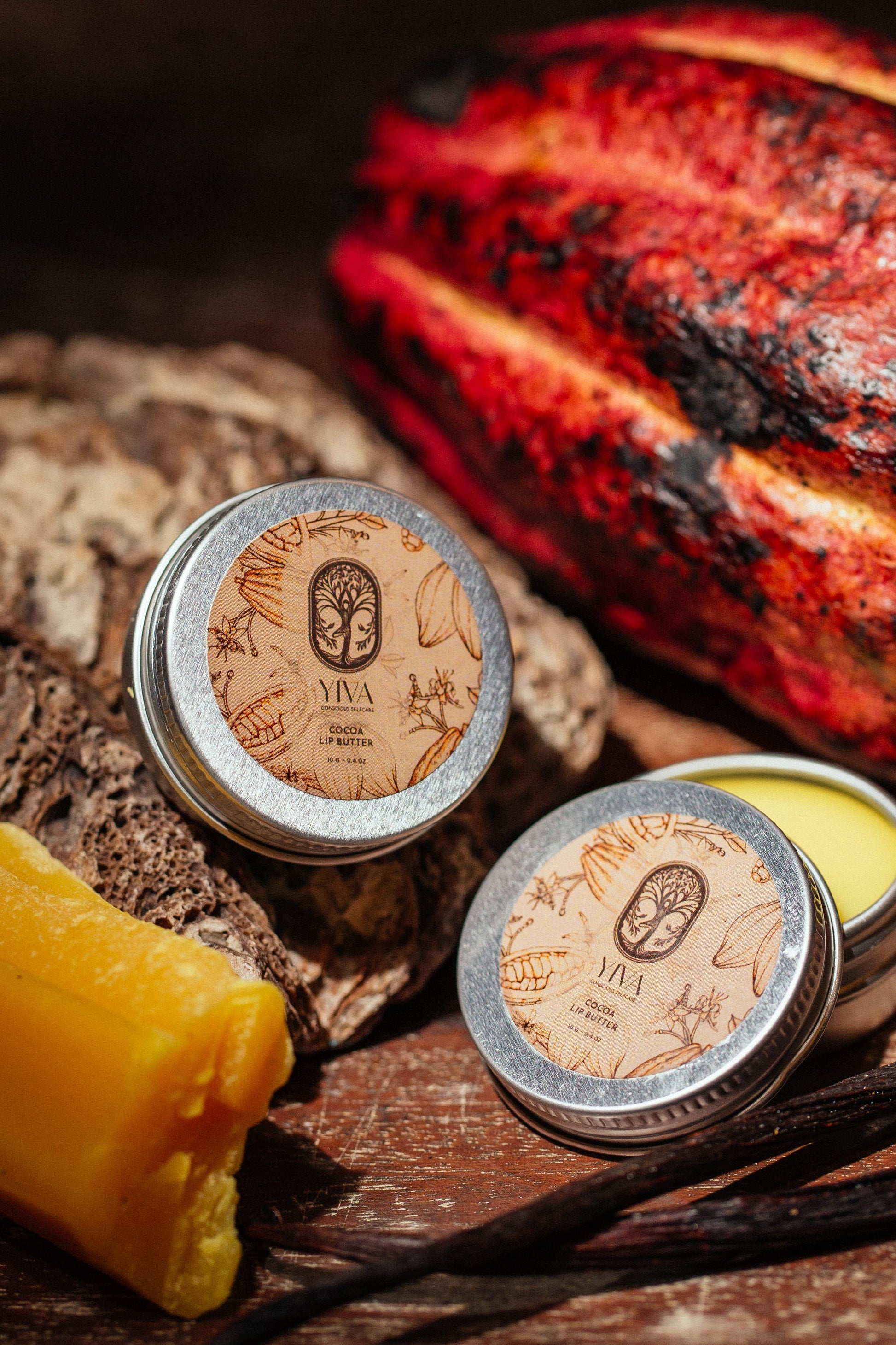 Two tins of Yiva Ceylon Lip Balm with a vanilla bean and cocoa pod on a wooden surface.