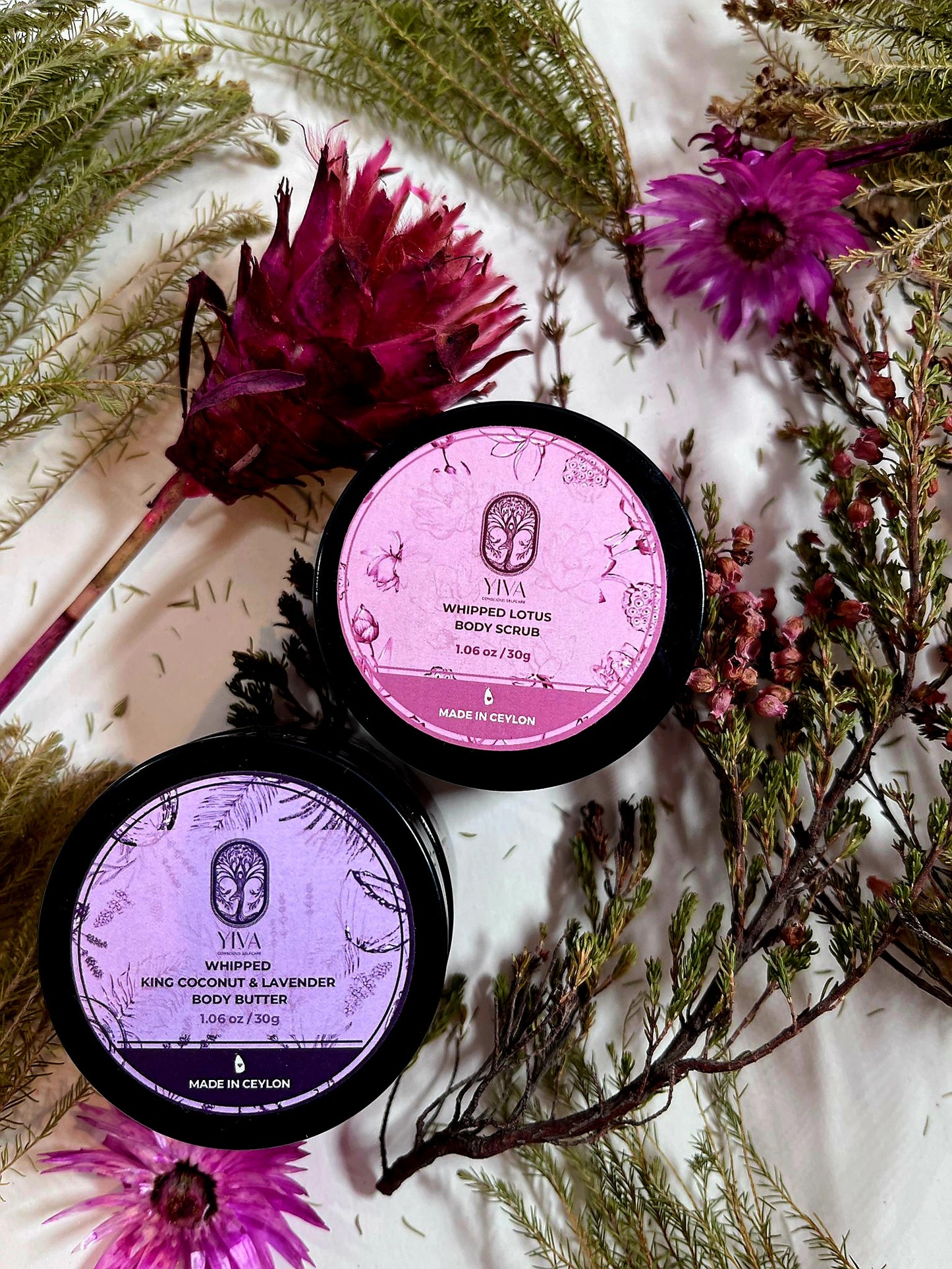 Two containers of Vya body butter on a white background with flowers and greenery.