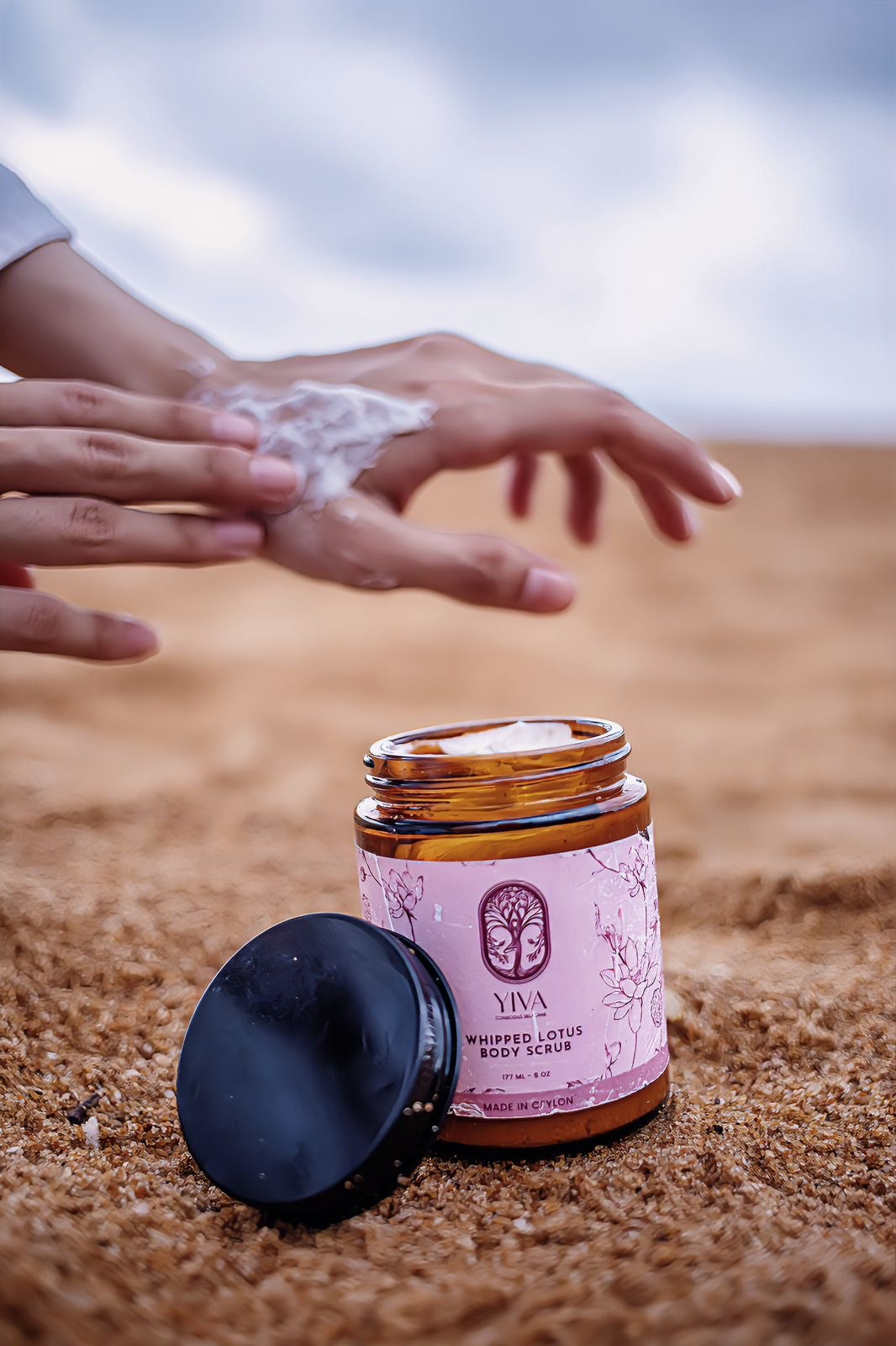Jar of VIVA Whipped Lotus Body Scrub on sand with hands applying product